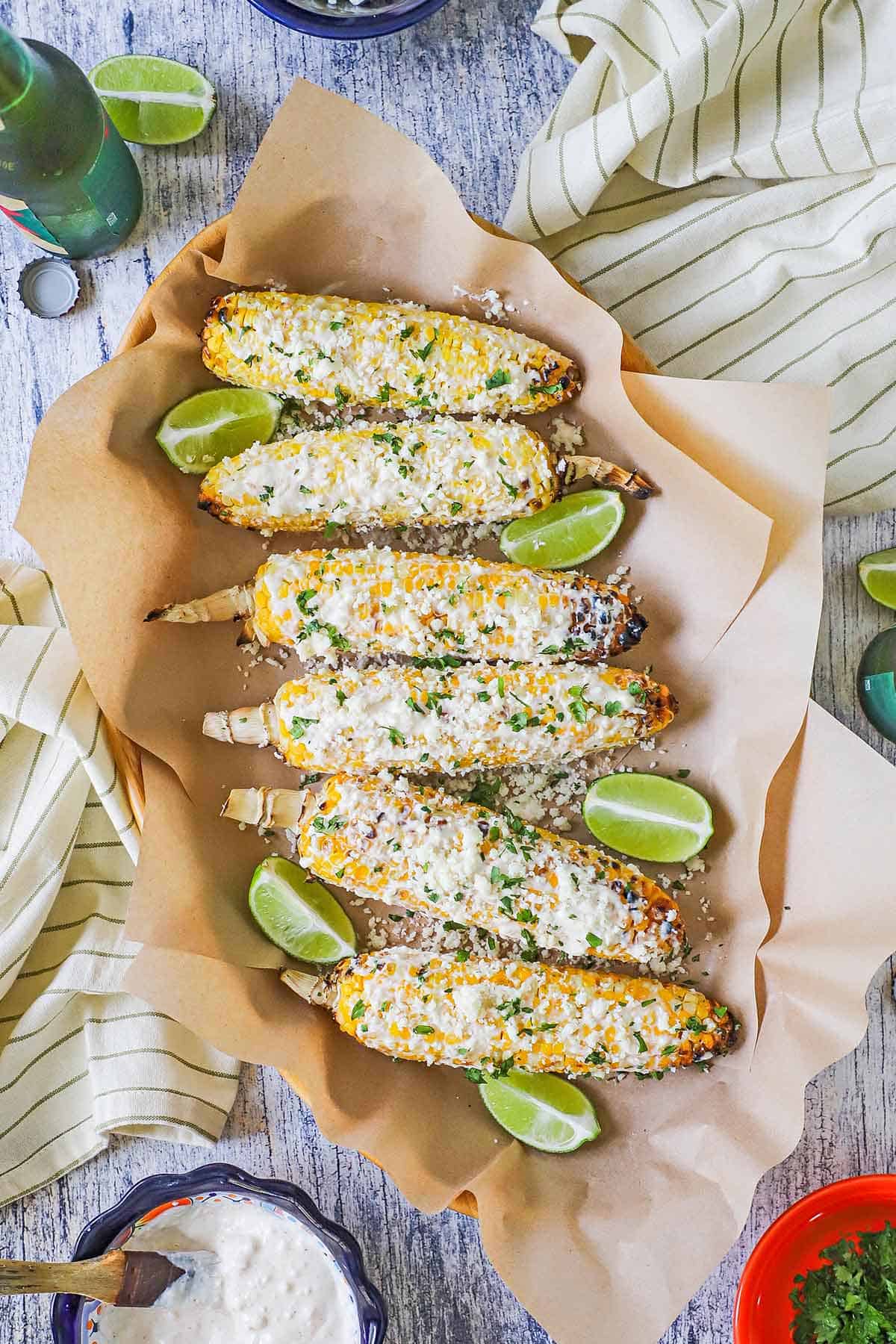A row six ears of Mexican street corn, eloté, resting on sheets of brown paper with lime wedges scattered around them.