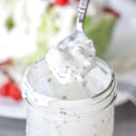A spoon filled with homemade blue cheese dressing being held over a jar filled with the dressing.