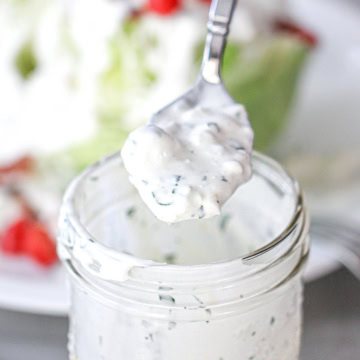 A spoon filled with homemade blue cheese dressing being held over a jar filled with the dressing.
