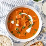 A white serving bowl filled with paneer butter masala and topped with streaks of cream and garnished with chopped cilantro.