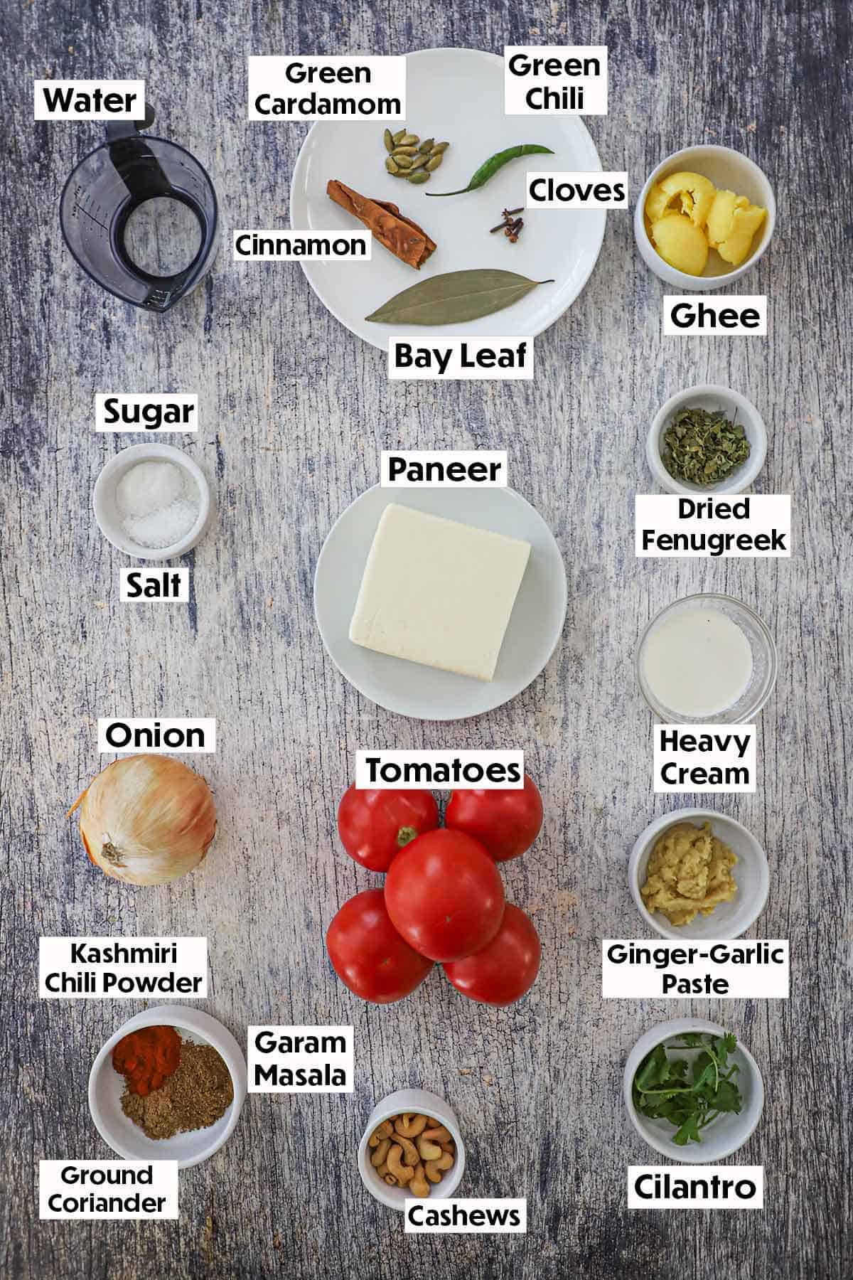 An arrangement of ingredients for paneer butter masala on a wooden grey background including a block of panner, tomatoes, an onion, seasonings, garlic-ginger paste, cream, and cilantro.