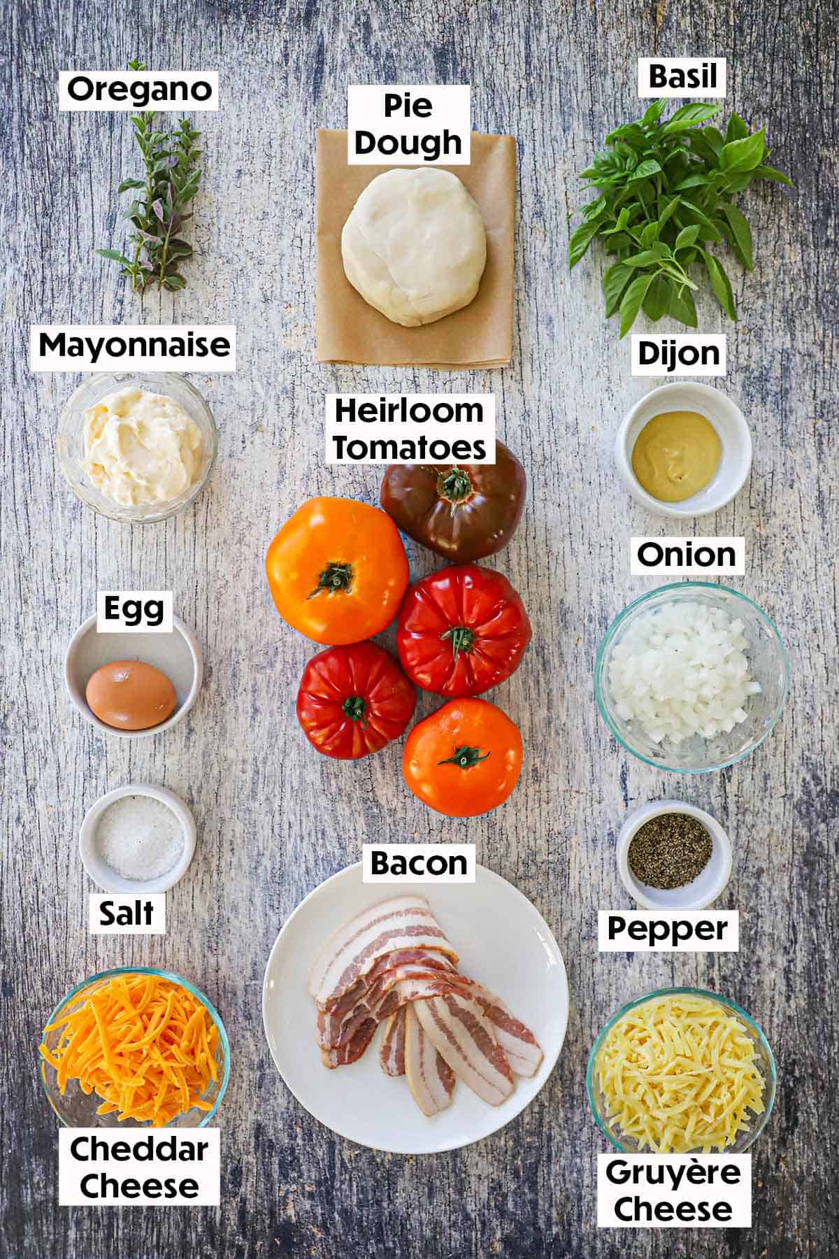An arrangement of ingredients for Southern tomato pie on a grey wooden background including heirloom tomatoes, pie dough, fresh herbs, mayonnaise, Dijon, onion, bacon, an egg, shredded cheese, salt, and pepper.