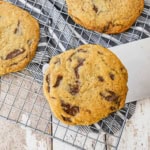 A large metal spatula being used to place a bakery-style chocolate chip cookie onto a baking rack with other cookies already on the rack.
