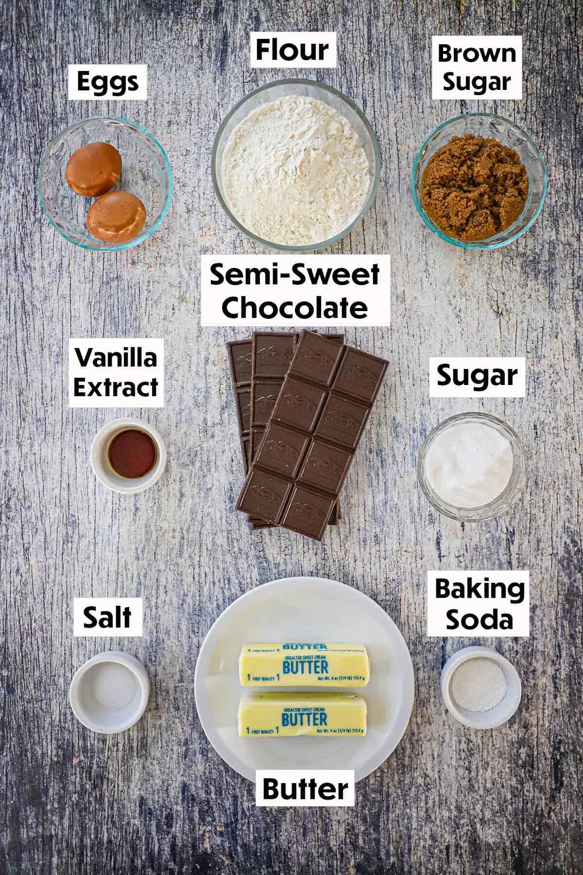 An arrangement of ingredients for bakery-style chocolate chip cookies on a grey wooden background including semi-sweet chocolate, eggs, flour, sugar, vanilla extract, salt, baking soda, and two sticks of butter.