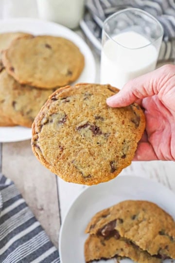 A person holding a large bakery-style chocolate cookie over a couple of plates of the cookies next to a tall glass of milk.