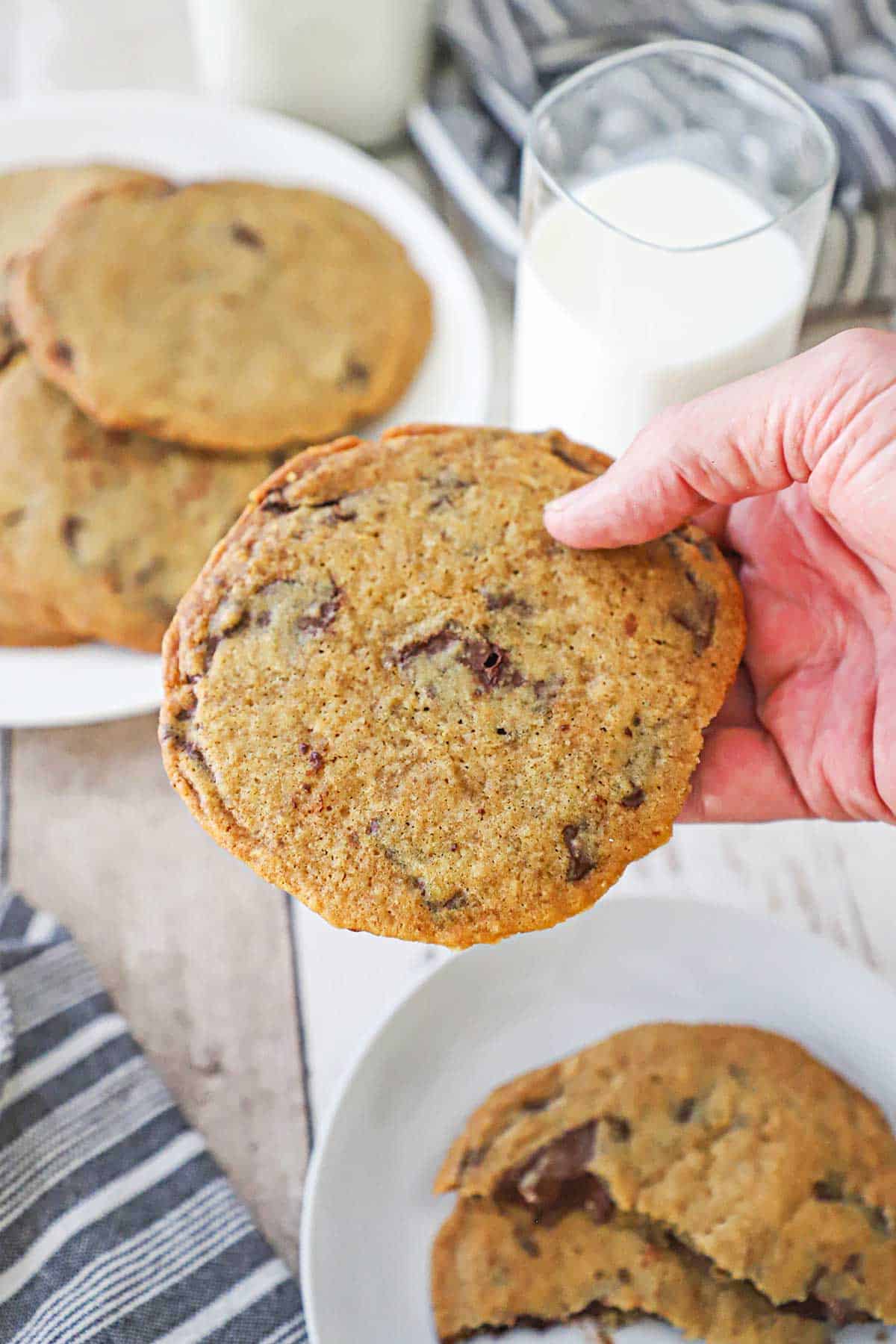 A person holding a large bakery-style chocolate cookie over a couple of plates of the cookies next to a tall glass of milk.