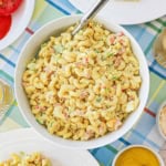 A white serving bowl filled with classic macaroni salad with a silver spoon inserted into the salad.