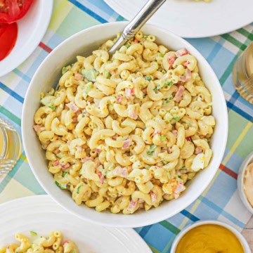A white serving bowl filled with classic macaroni salad with a silver spoon inserted into the salad.
