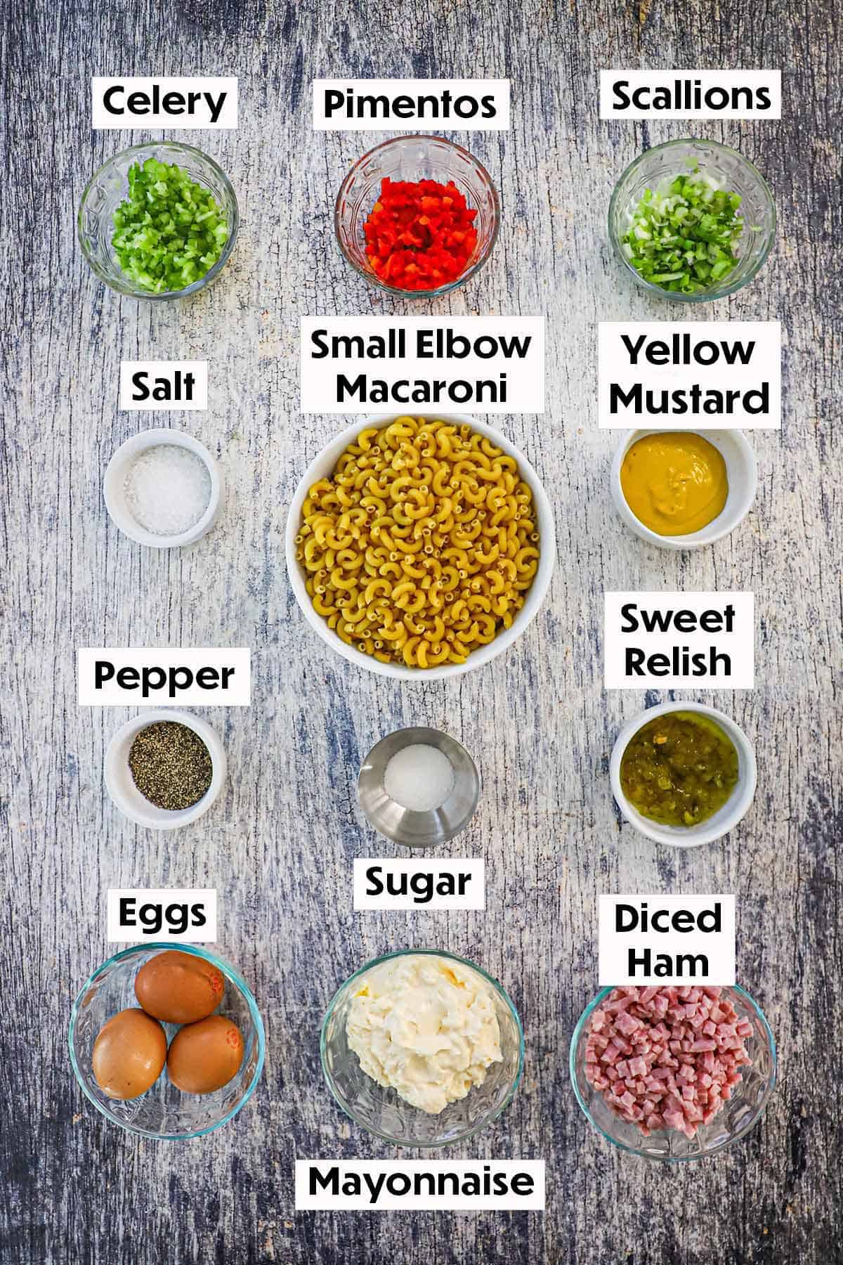 An arrangement of ingredients for classic macaroni salad on a wooden grey background including dried macaroni, celery, pimentos, scallions, mustard, relish, eggs, mayonnaise, diced ham, and seasonings.