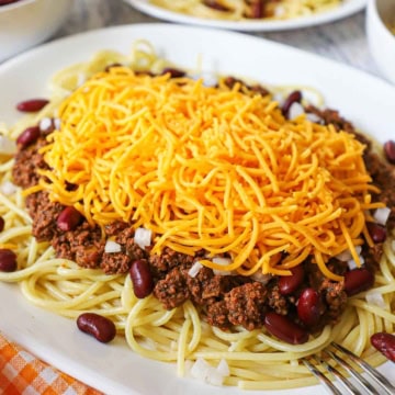 An oval white plate filled with Cincinnati chili served 5 ways including the chili over cooked spaghetti along with red kidney beans, chopped onion, and topped with a large mound of finely shredded cheddar cheese.