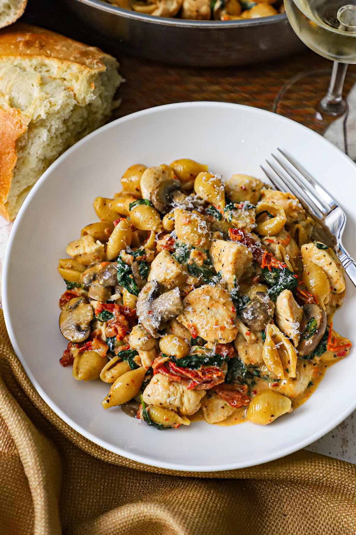 A white bowl filled with a serving of One-Pot Marry Me Chicken Pasta topped with shaved Parmesan cheese with a torn piece of Italian bread nearby.