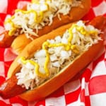A close-up view of two Coney Island hot dogs that are topped with sauerkraut, chopped onions, and a zig zag of deli-style mustard.