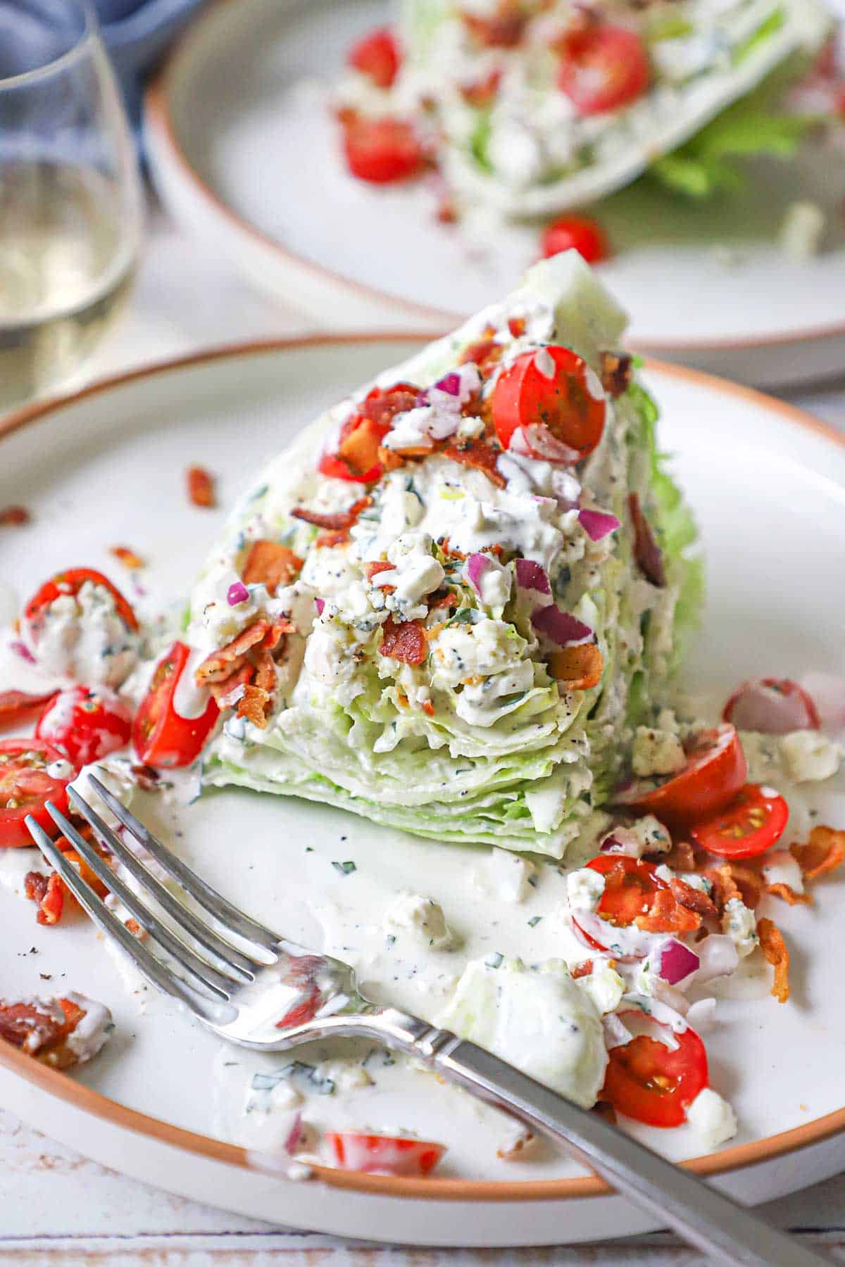A partially eaten steakhouse wedge salad on a white plate with iceberg lettuce, cherry tomatoes, crumbled bacon, and blue cheese dressing.