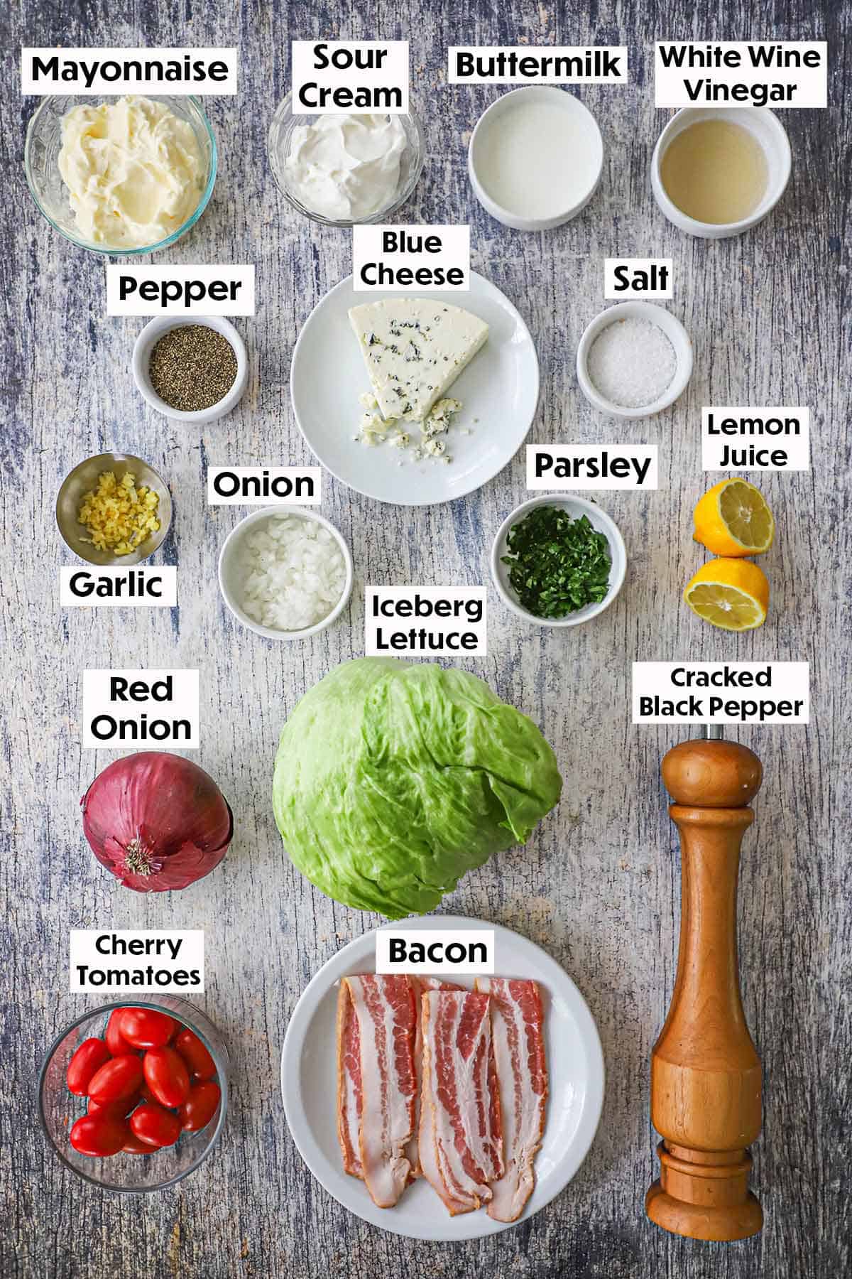 An arrangement of ingredients for steakhouse wedge salad on a grey wooden background including everything for homemade blue cheese, as well as a red onion, iceberg lettuce, cherry tomatoes, uncooked bacon, and a pepper grinder.