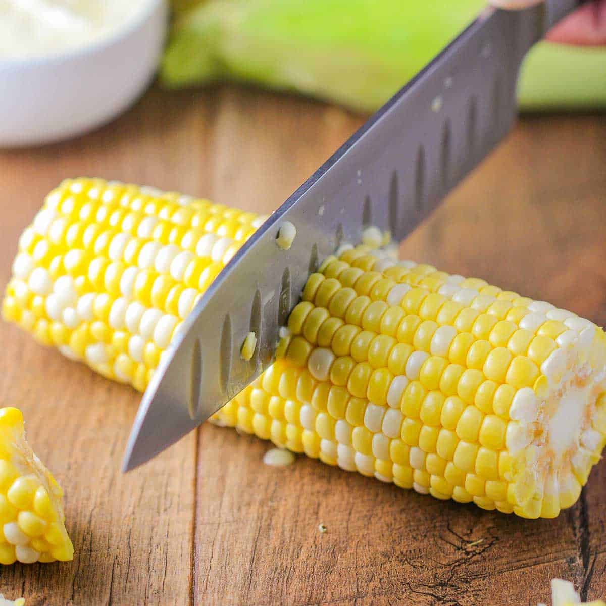 A person using a large chef's knife to cut a freshly shucked ear corn in half on a wooden cutting board.
