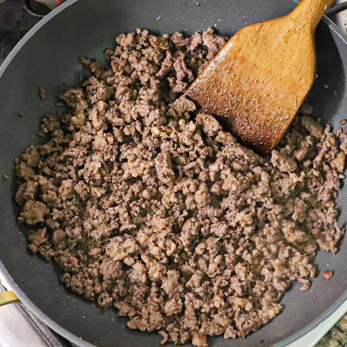 A non-stick skillet filled with fully cooked ground beef and Italian sausage with a wooden spatula off to the side.