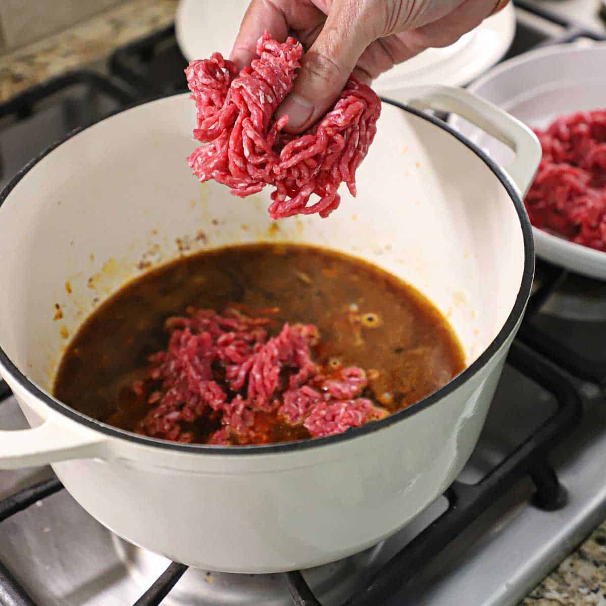 A person using his fingers to crumble ground beef into a white pot filled with a brown simmering sauce.