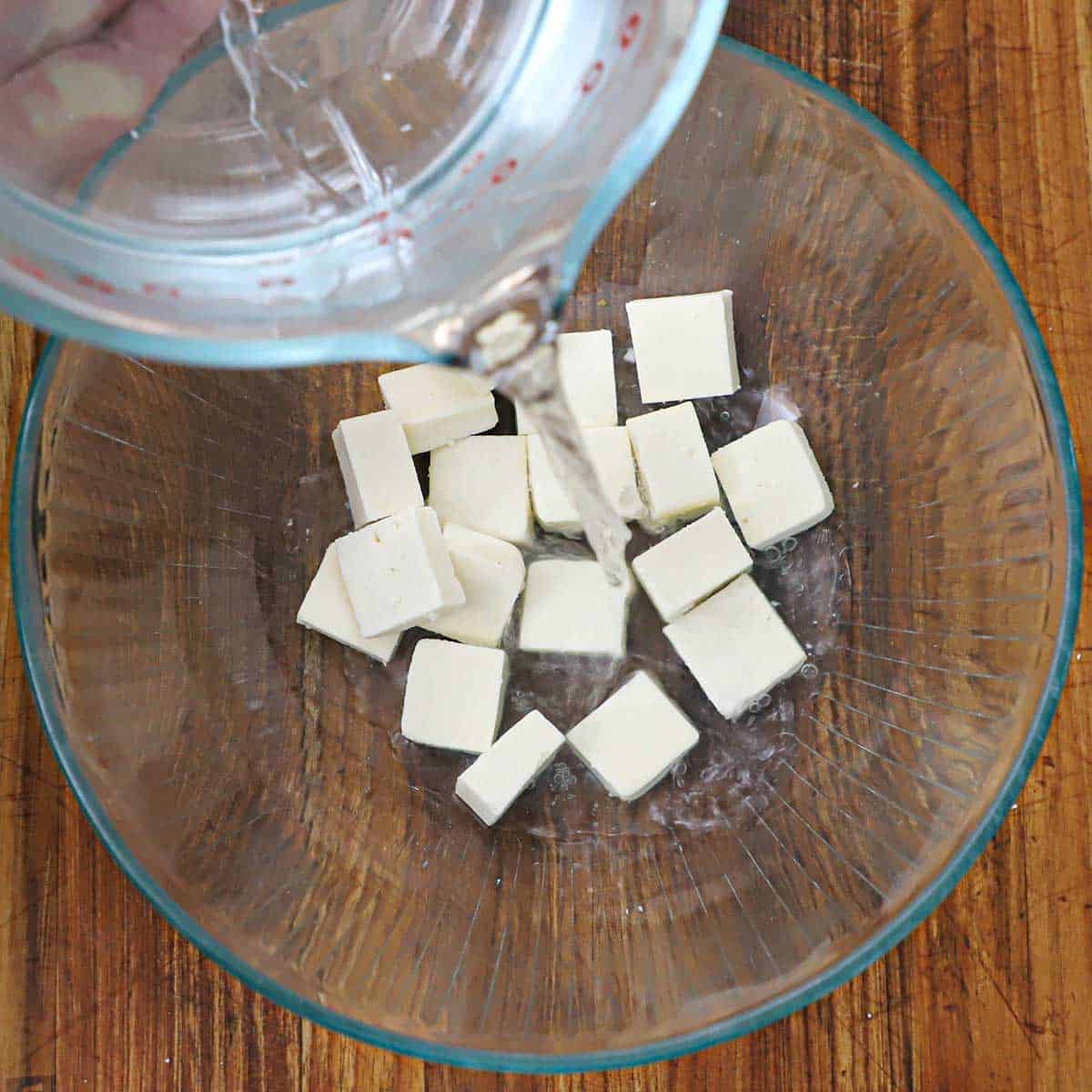A person pouring warm water from a large glass measuring cup into a glass bowl that has a layer of bite-sized paneer in the bottom of it.