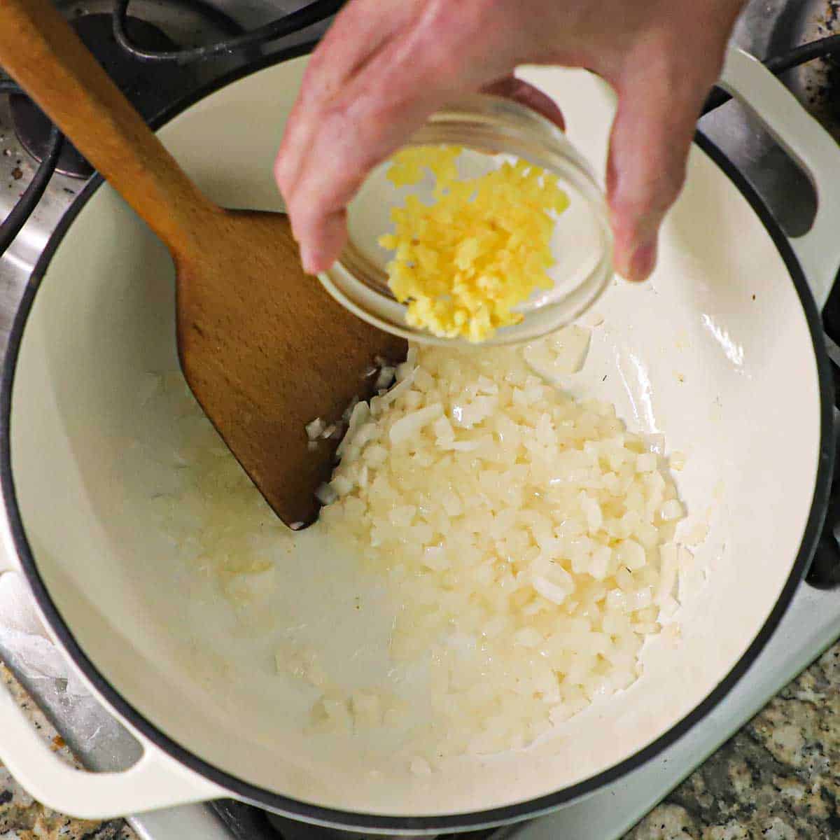 A person dumping minced garlic from a small glass bowl into a white pot filled with simmering chopped onions on a gas stove.