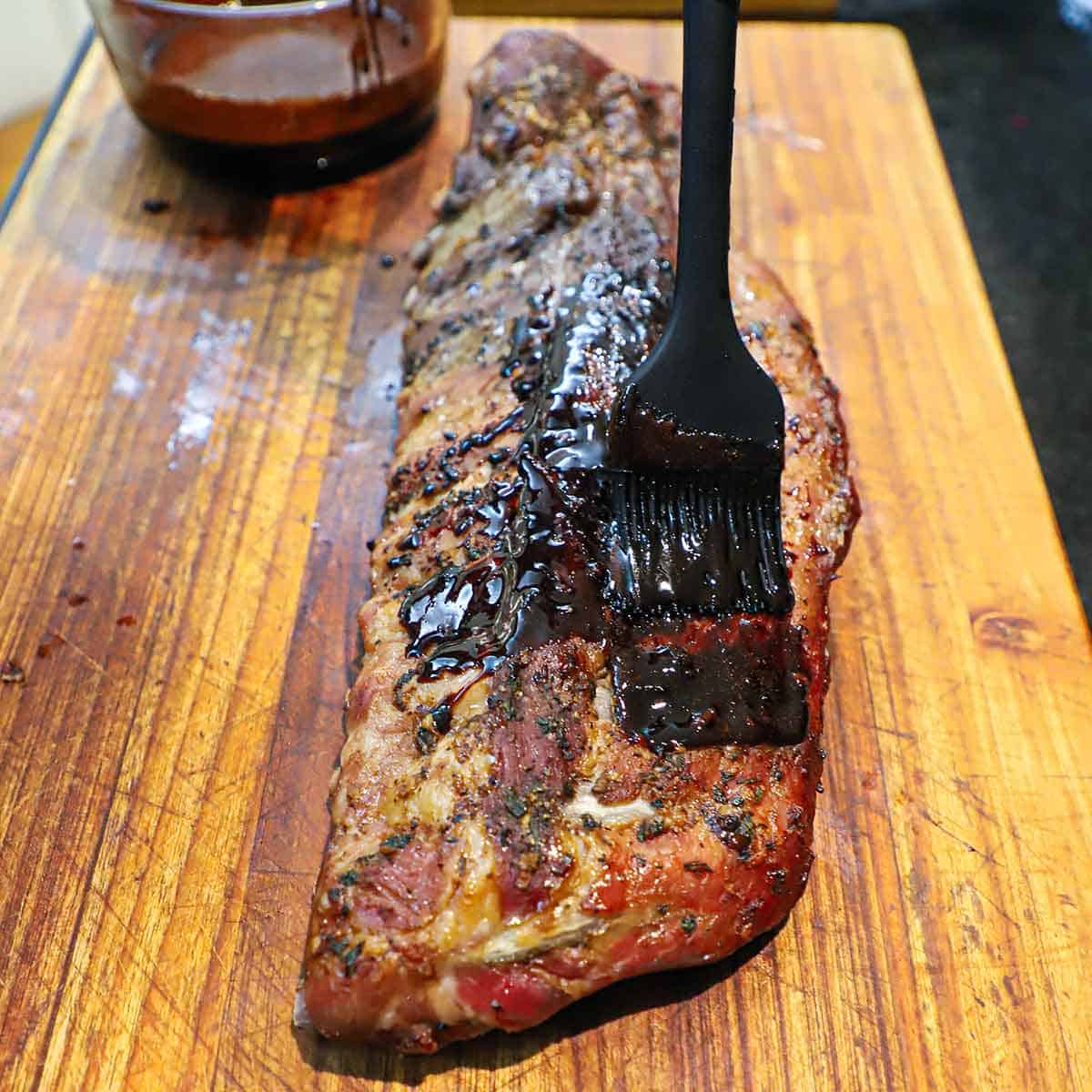 A person using a grill brush to apply a balsamic glaze over the surface over a rack of oven-roasted baby back ribs.
