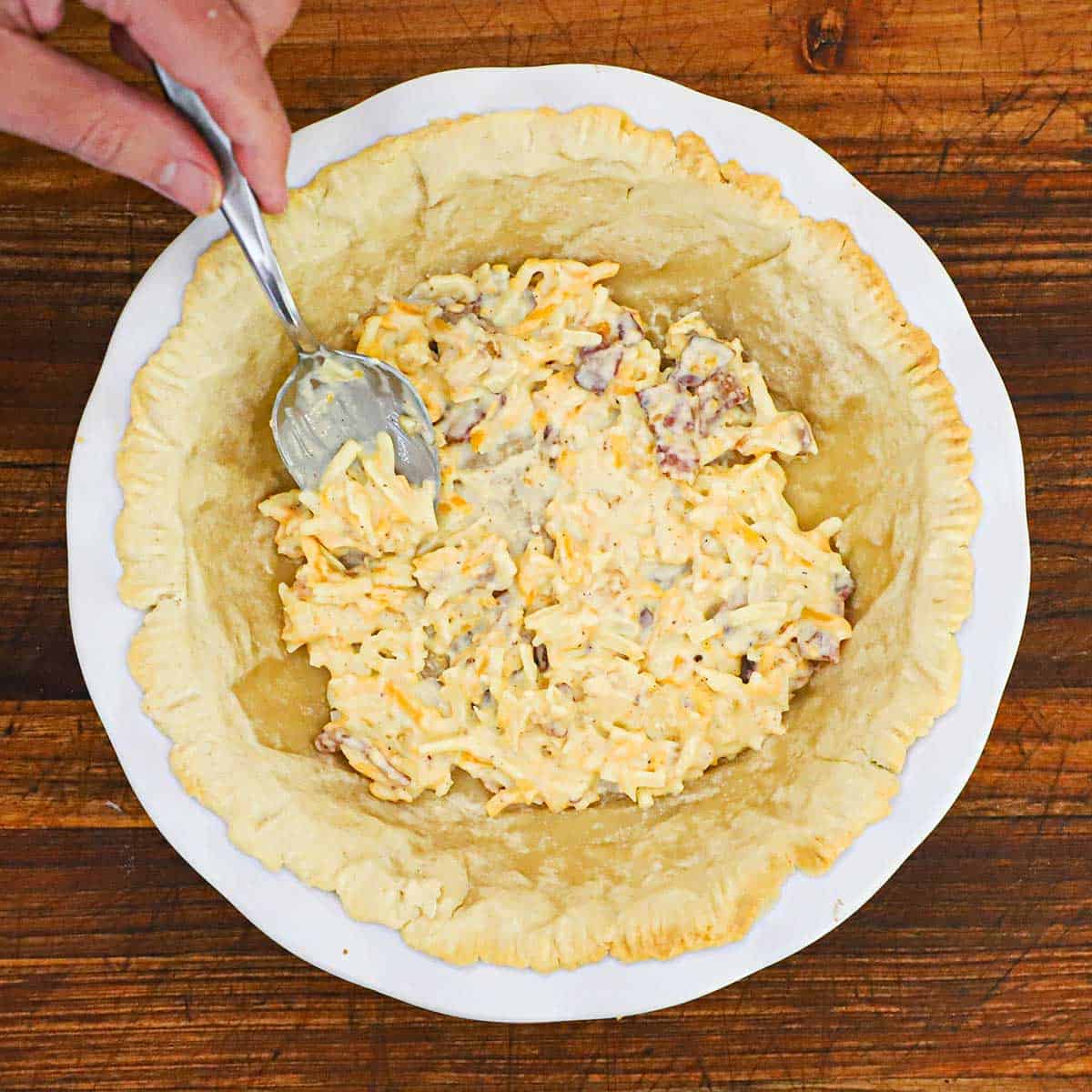 A person using a spoon to add a cheese, bacon, and mayonnaise mixture into the bottom of a pie dish lined with a partially baked pie crust.