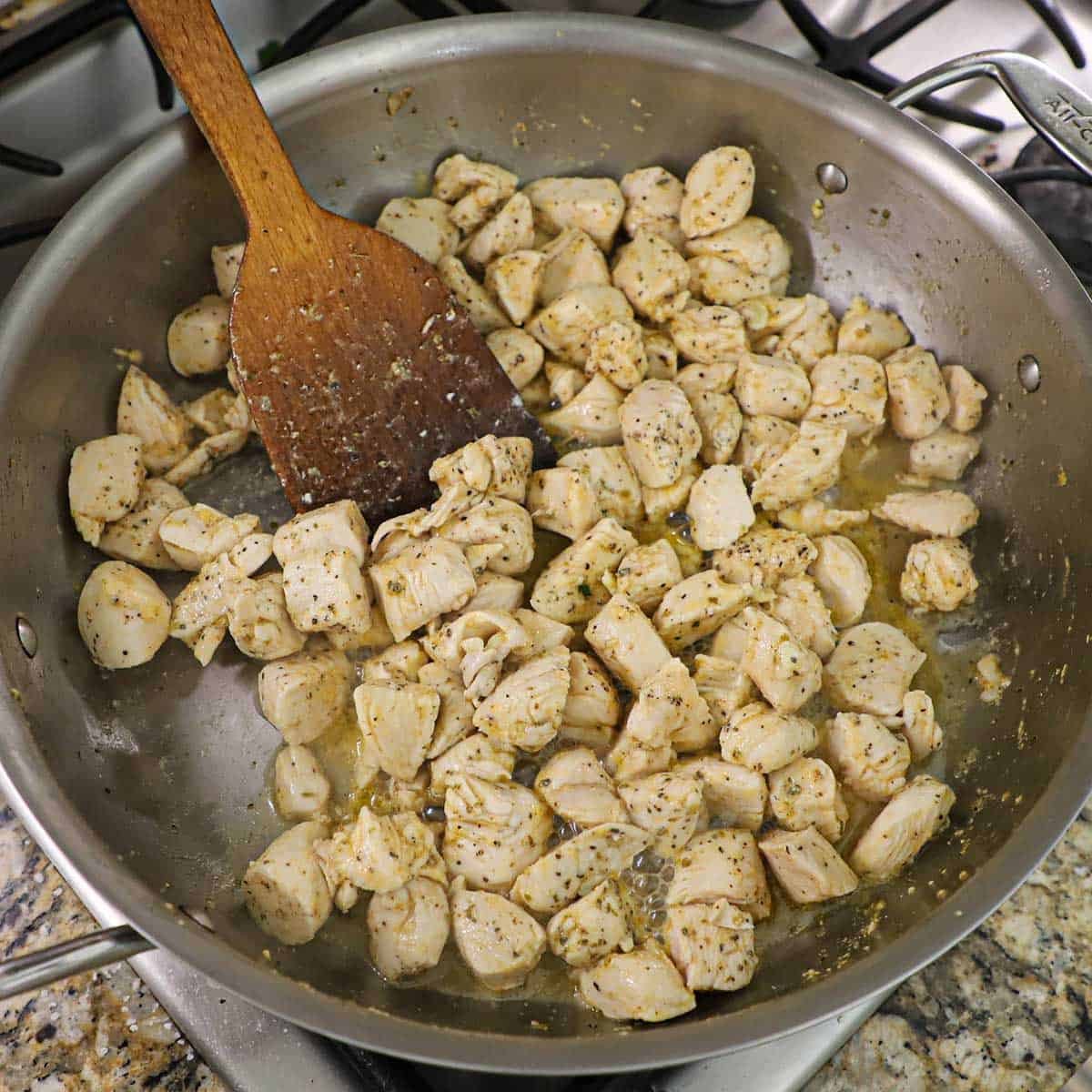 A person cooking bite-size pieces of chicken in a stainless steel skillet in a oil and herbs with a large wooden spatula inserted in the middle of the chicken.