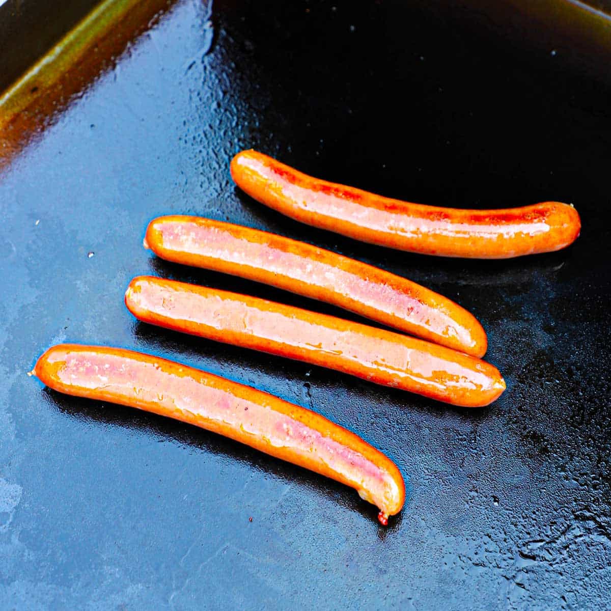 Four natural casing beef frankfurters being cooked on a large black griddle.