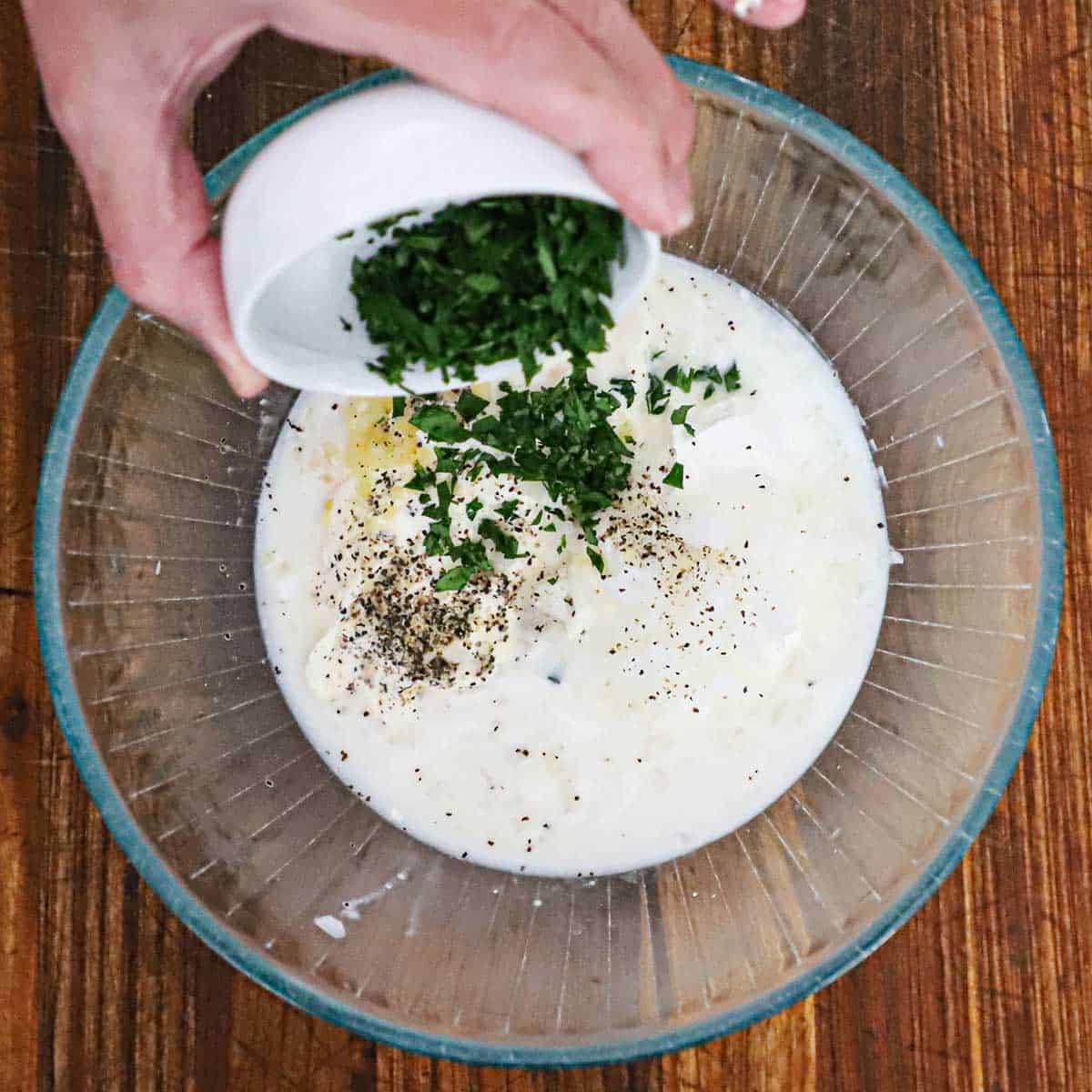 A person transferring chopped Italian parsley from a small white bowl into a glass bowl filled with mayonnaise, sour cream, buttermilk, lemon juice, and seasonings.