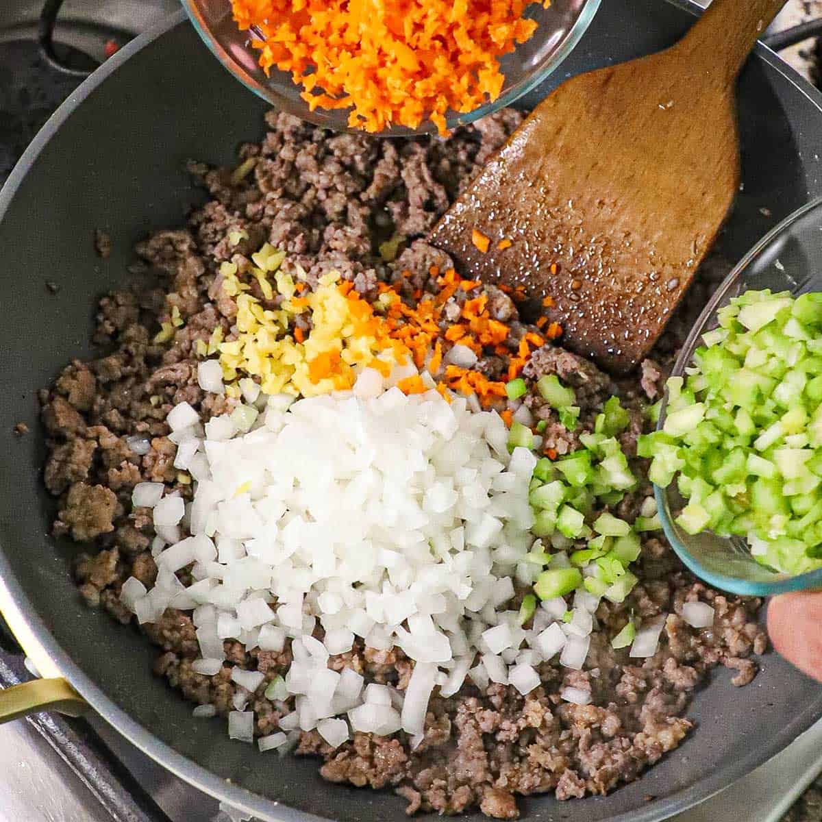A person dumping chopped carrots from a small bowl with one hand and another bowl filled with chopped celery both going into a skillet filled with cooked beef, sausage, as well as chopped onions and minced garlic.