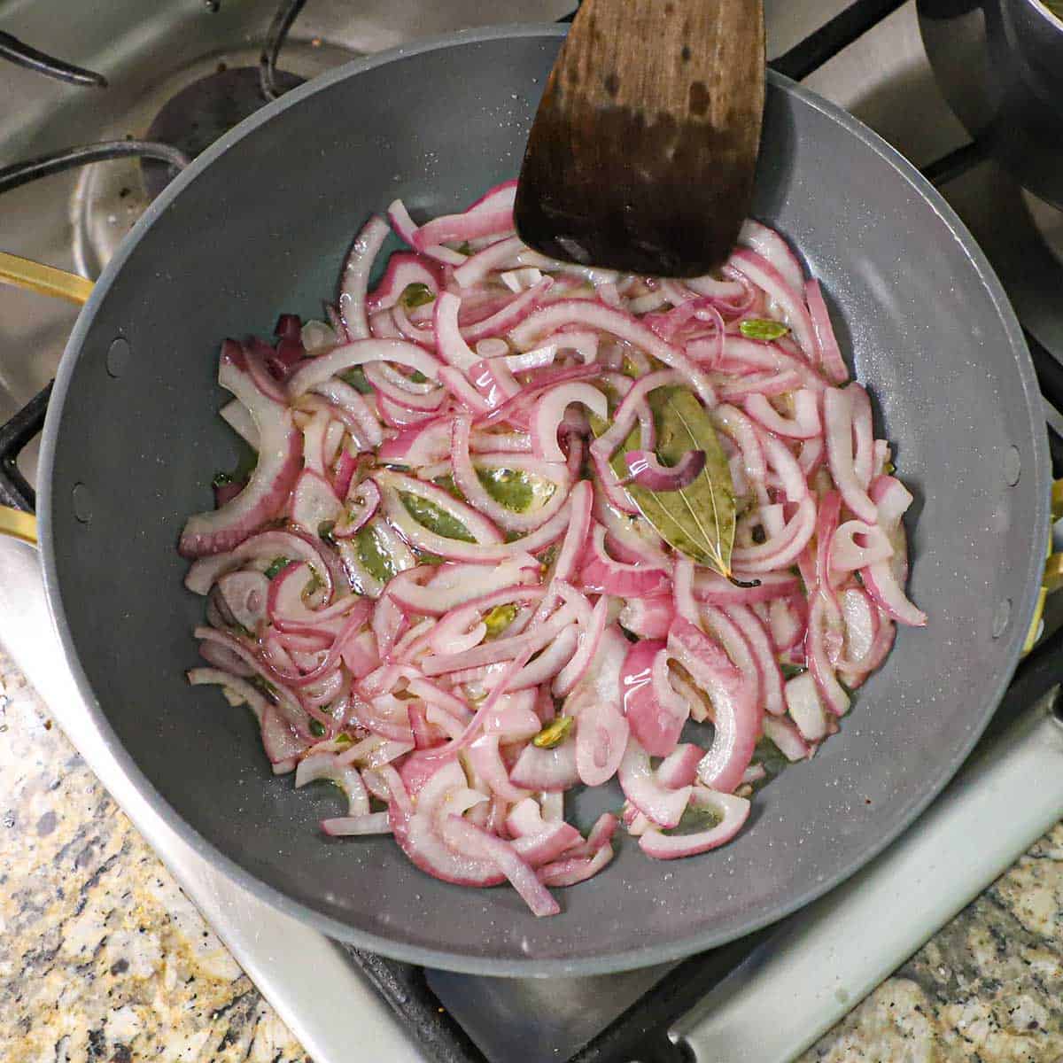 A large non-stick skillet filled with strips of red onion that are being sautéed in ghee along with a large Indian bay leaf and green cardamom pods.