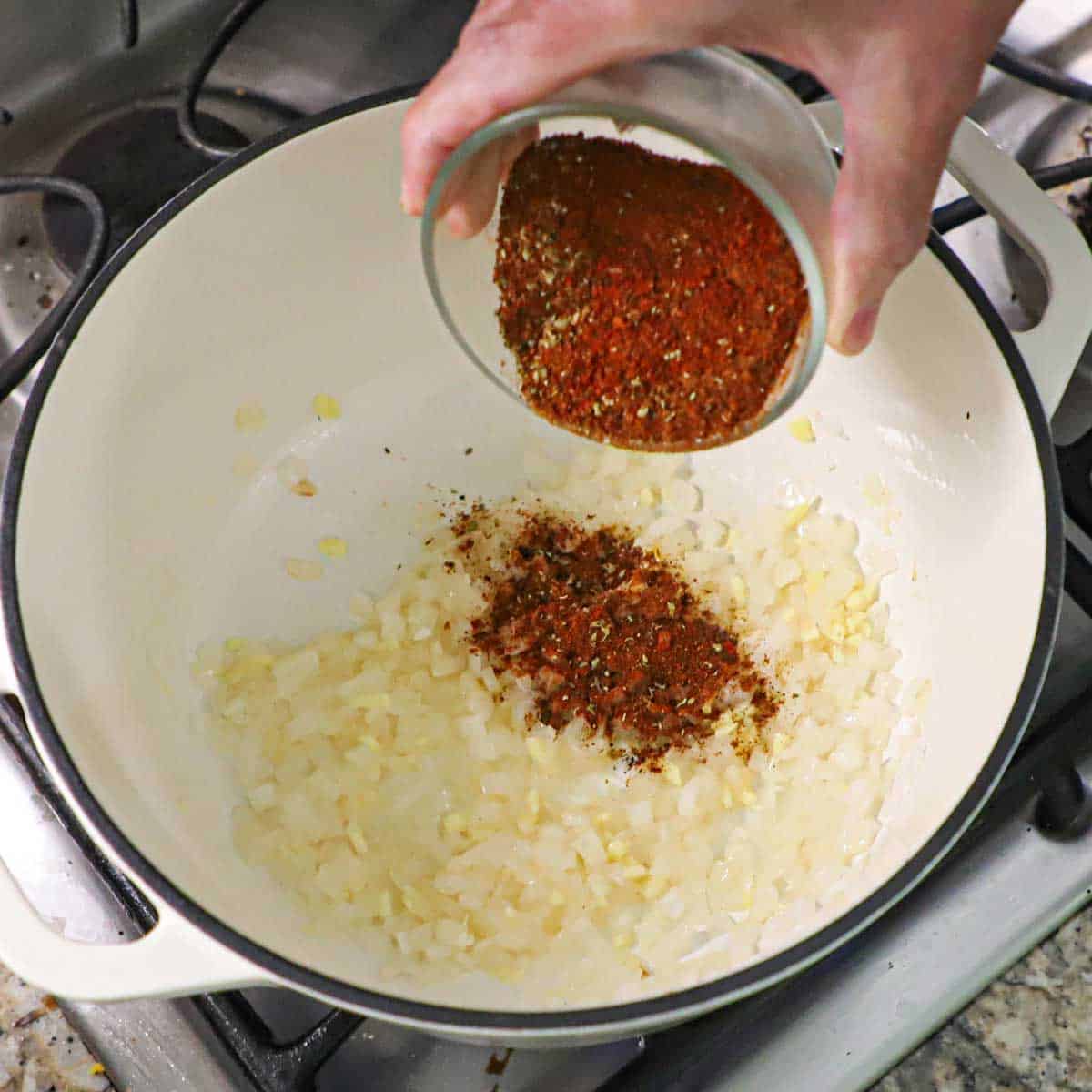 A person transferring a combination of seasonings for Cincinnati chili into a pot filled with sautéed onion and garlic.