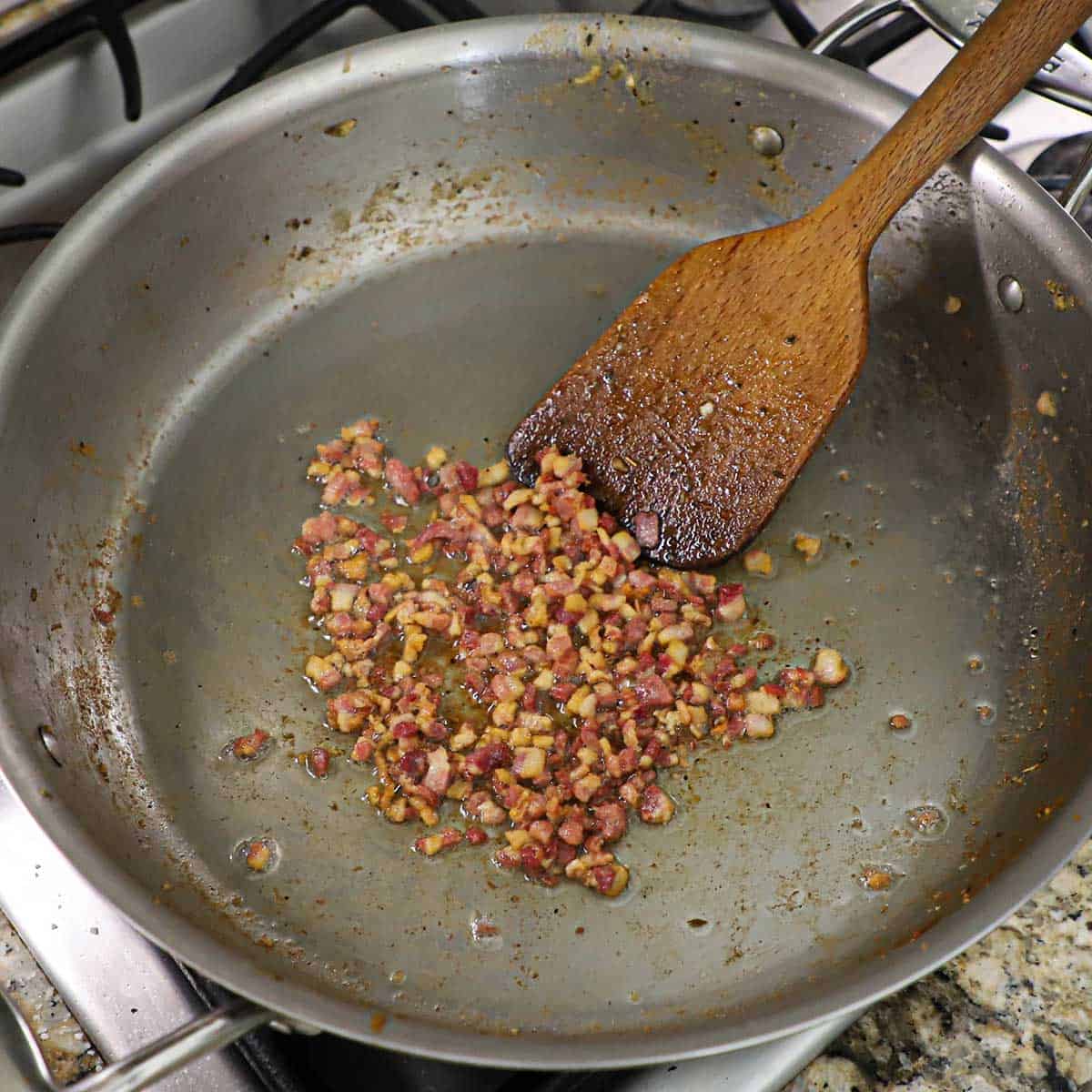 A large wooden spatula being used to stir crispy cubed pancetta that has been cooked in a large silver skillet.