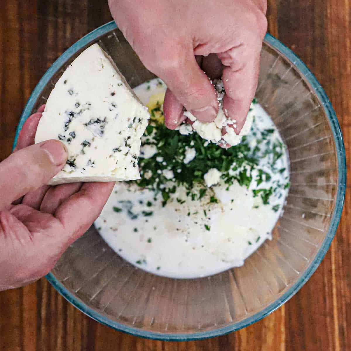 A person using his fingers to crumble a block of blue cheese into a glass bowl filled with mayonnaise, buttermilk, sour cream, herbs, and seasonings.
