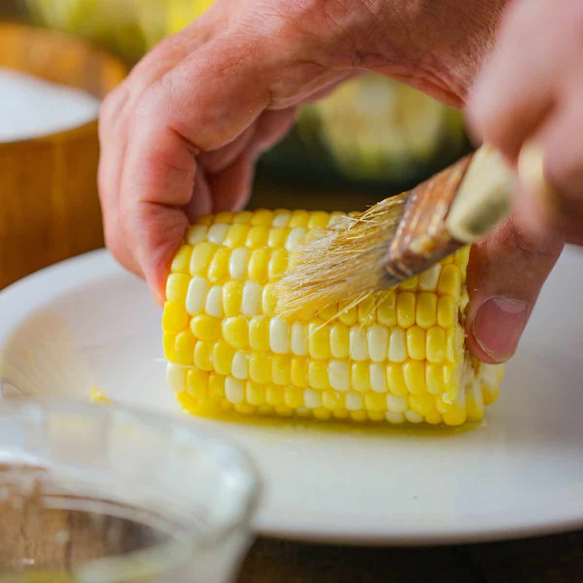 A person holding a shucked corn cob that has been cut in half and is being held on a white plate while the other hand brush butter on it.
