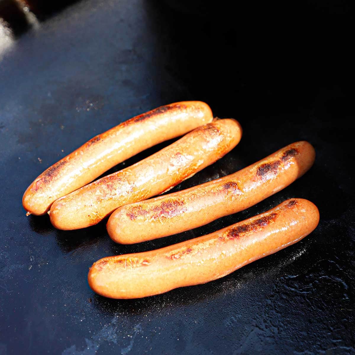 Four all-beef natural casing hot dogs being cooked on a large outdoor griddle.