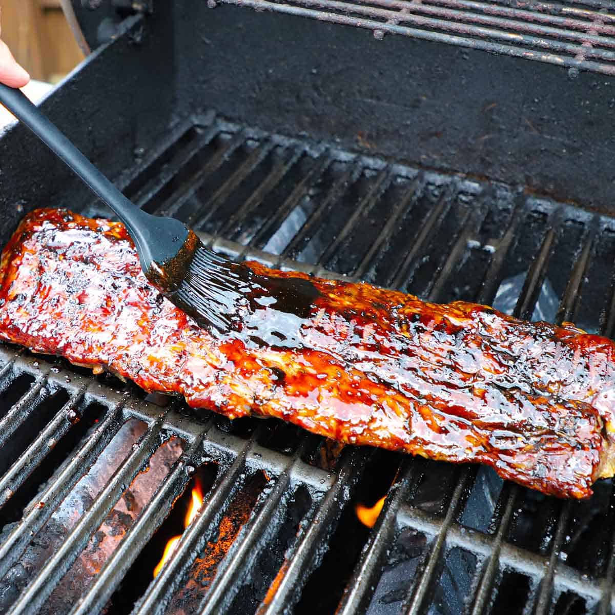 A person using a grill brush to apply a balsamic glaze onto the top side of a rack of baby back ribs being cooked on the grate of a gas grill.