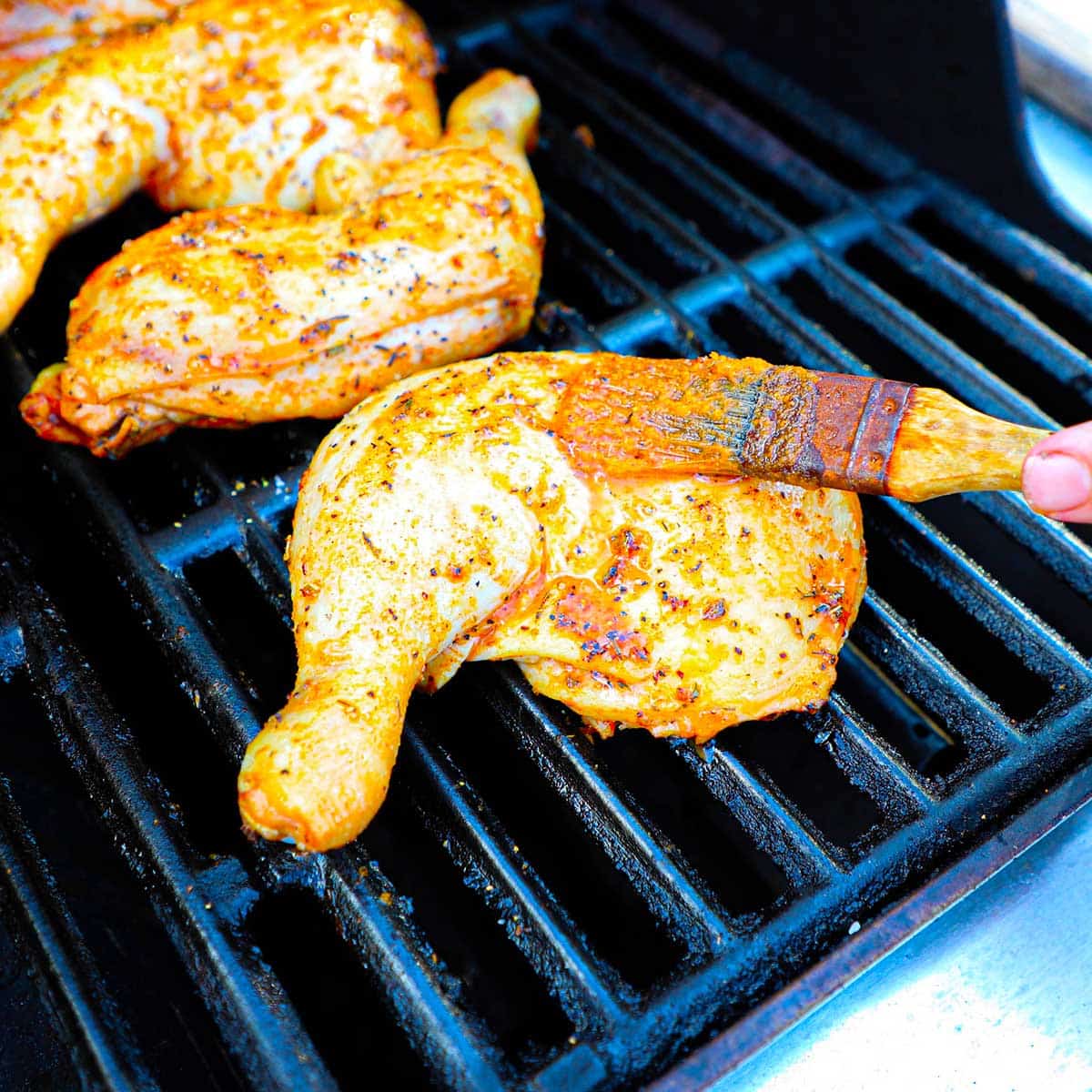 A person using a pastry brush to baste chicken leg quarters that are being grilled on a gas grill.