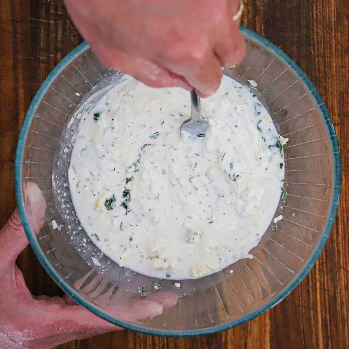 A person using a fork to mix together the ingredients for homemade blue cheese dressing in a glass bowl on a wooden cutting board.