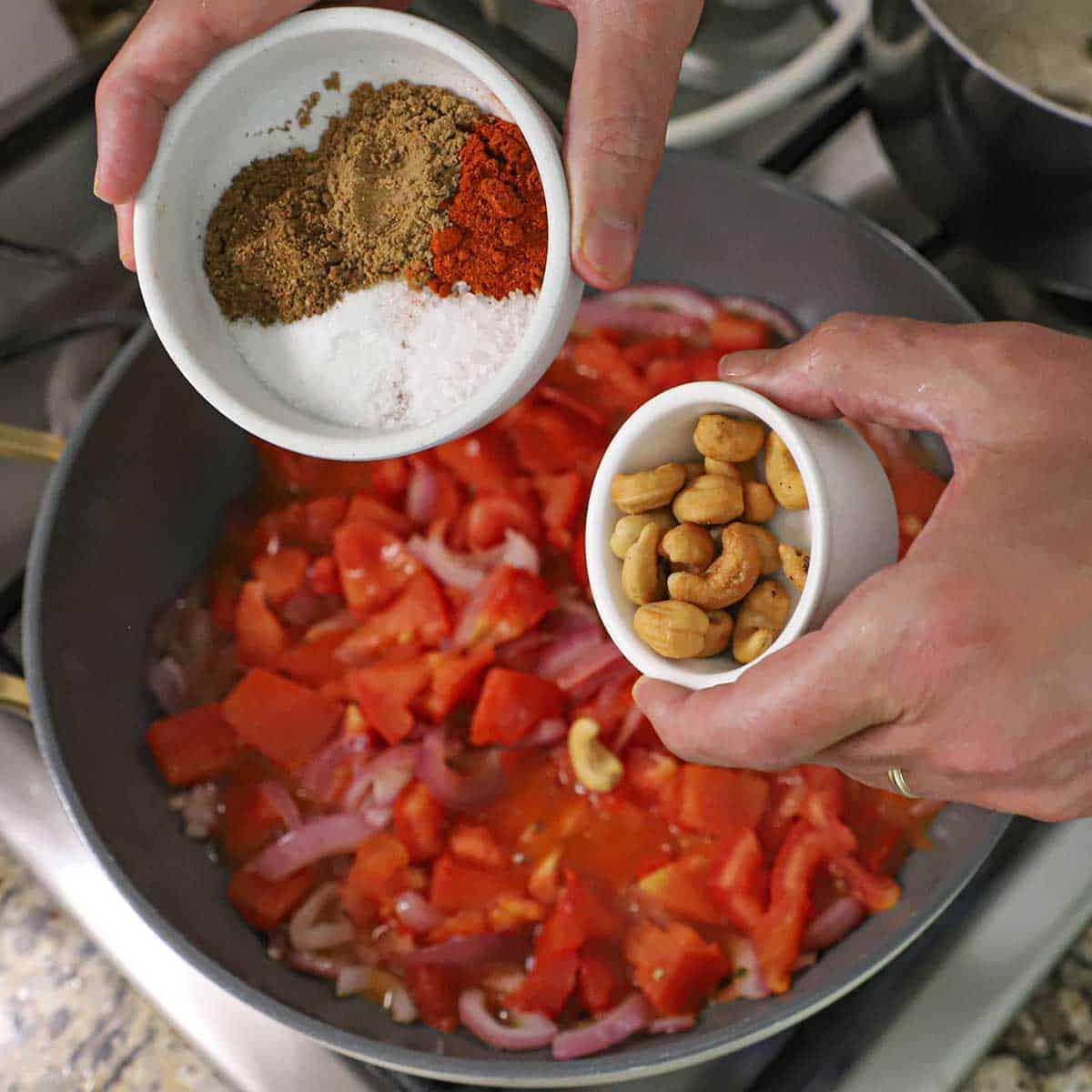 A person holding a small bowl of cashews in one hand and another bowl filled with salt, sugar, garam masala, coriander, and chili powder all over a skillet filled with simmering, tomatoes and red onions.