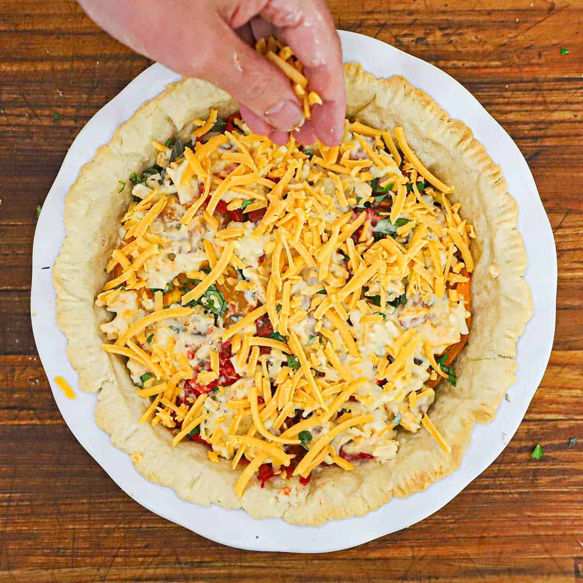 A person sprinkling shredded cheddar cheese over the top of an un-baked Southern tomato pie.