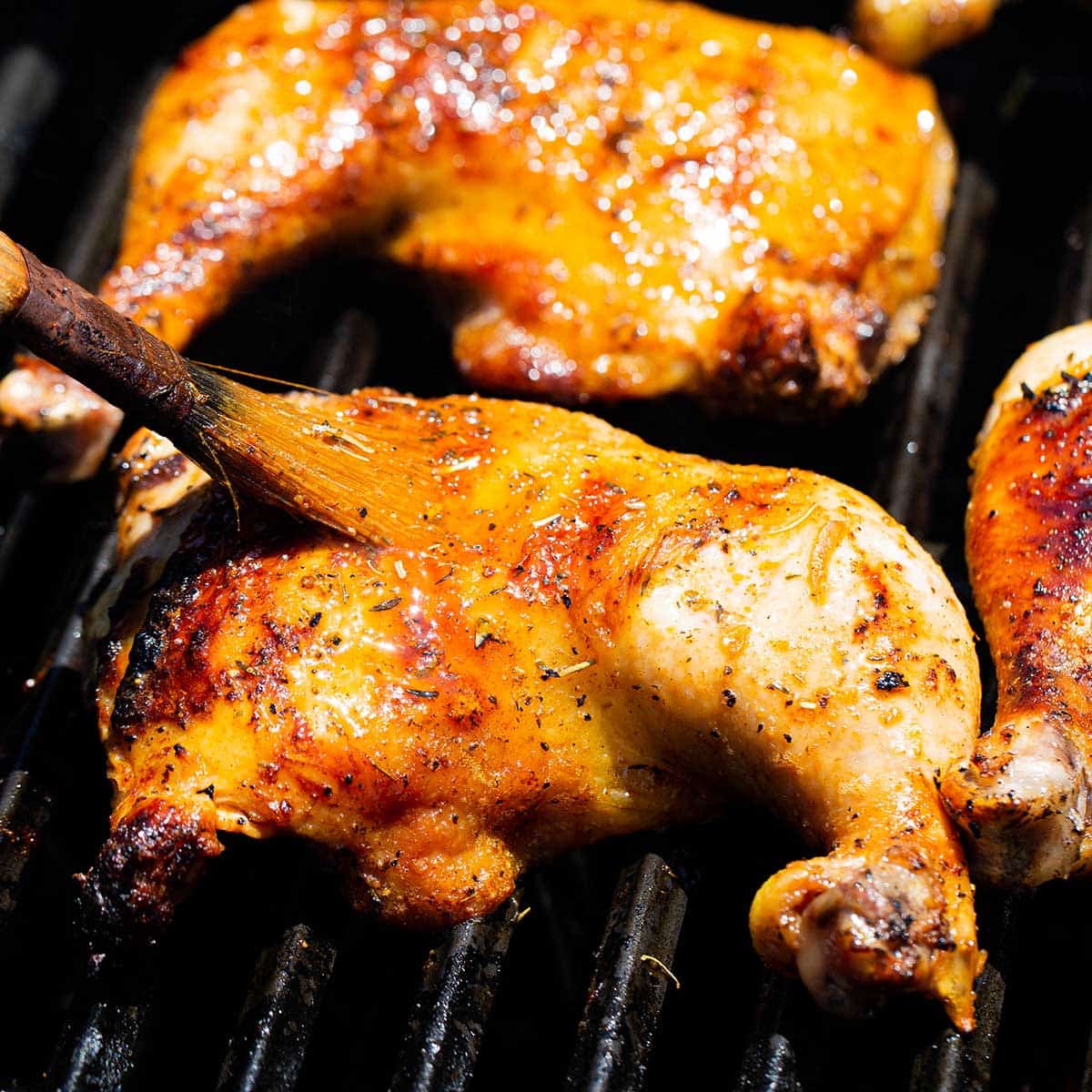 A person using a pastry brush to apply melted butter onto best grilled chicken leg quarters over non-direct heat on a gas grill.