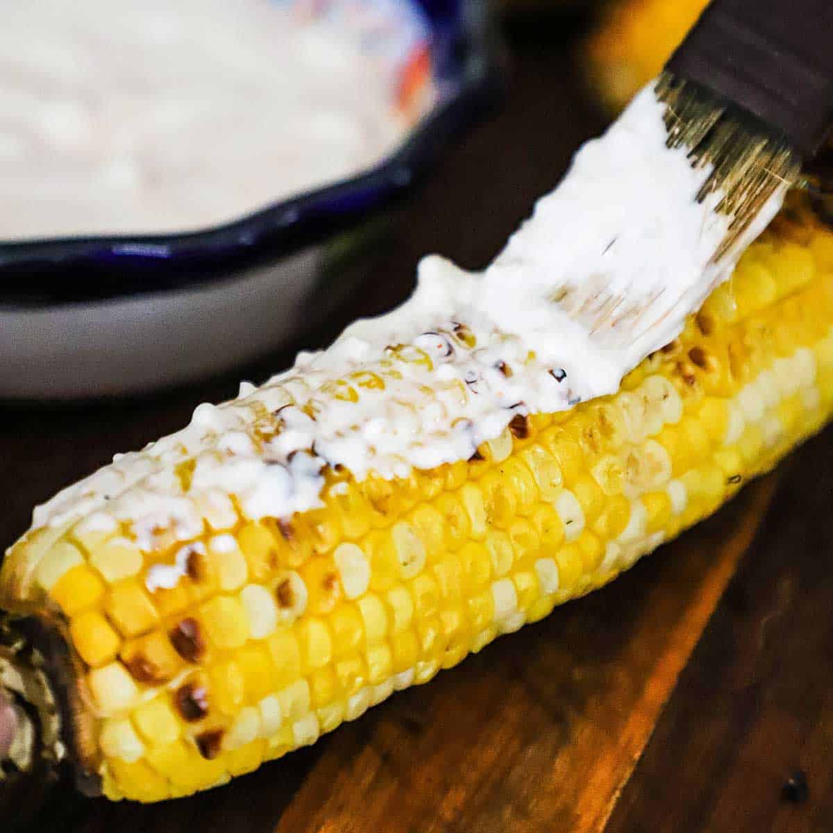 A person brushing a cream and cheese mixture over the surface of an ear of corn that has been grilled.