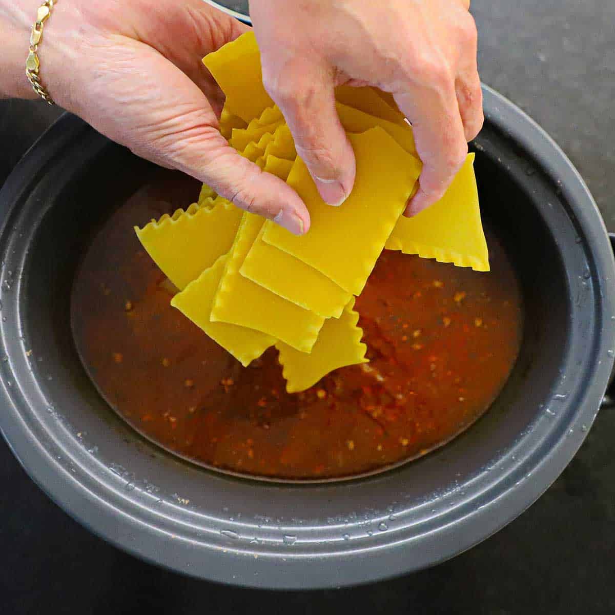A person dropping pieces of dried lasagna noodles into a slow cooker filled with lasagna soup.