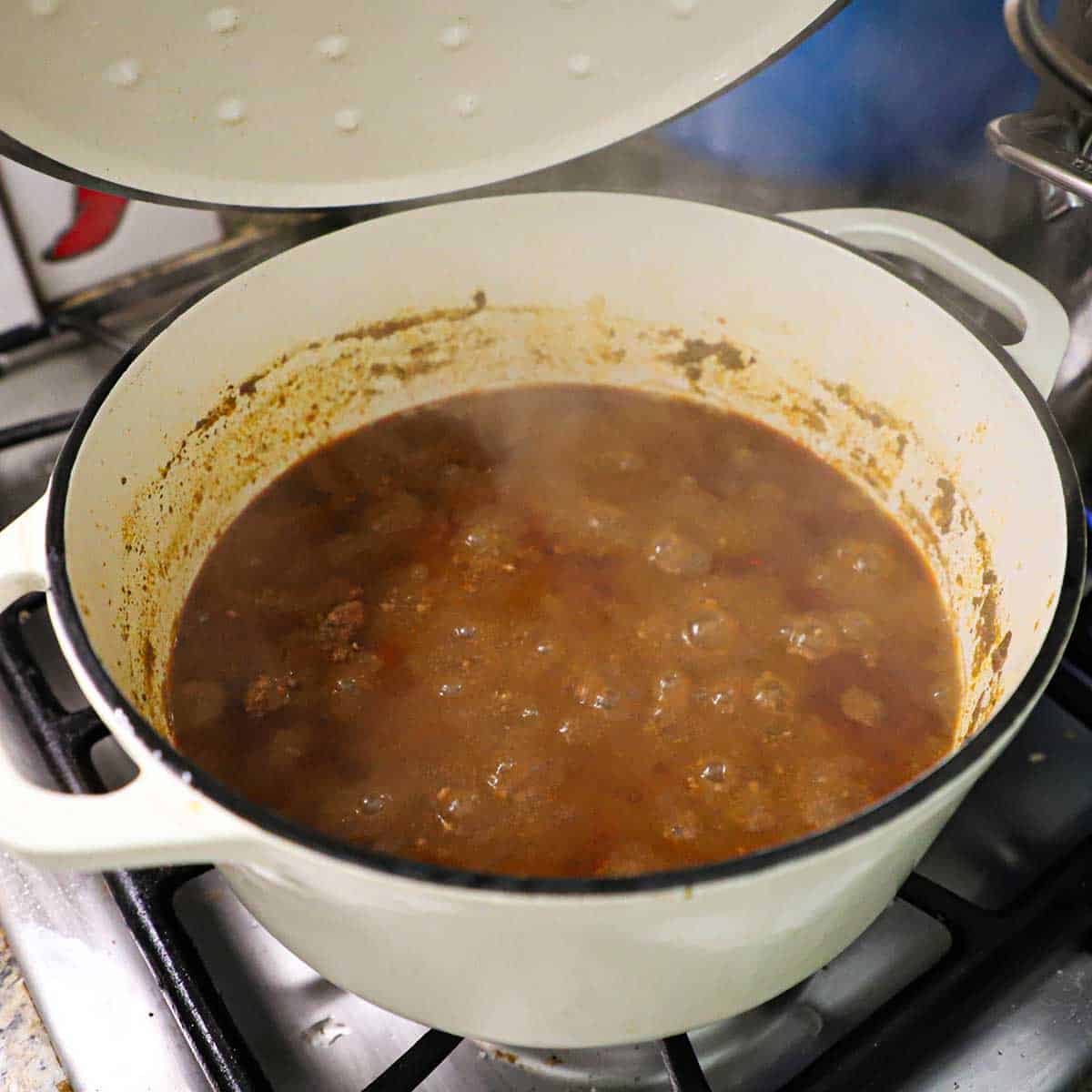 A person lifting a lid from a large white pot that is filled with simmering Cincinnati chili on a gas stove.