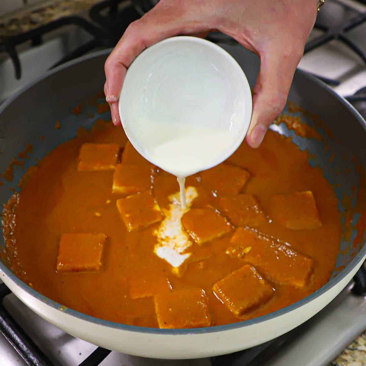 A person pouring heavy cream from a small white bowl into a skillet filled with simmering paneer butter masala.
