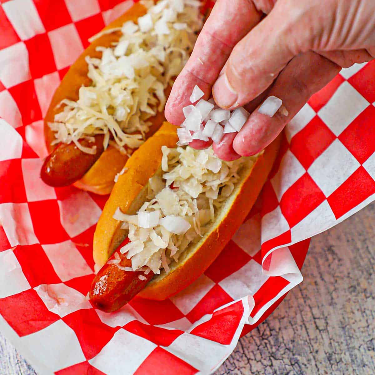A person sprinkling chopped onion over the top of a Coney Island hot dog resting in a basket lined with checkered wax paper.