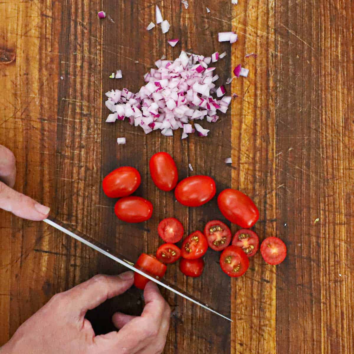 A person cutting cherry tomatoes into slices on a wooden cutting board with a pile of chopped red onion next to the tomatoes.