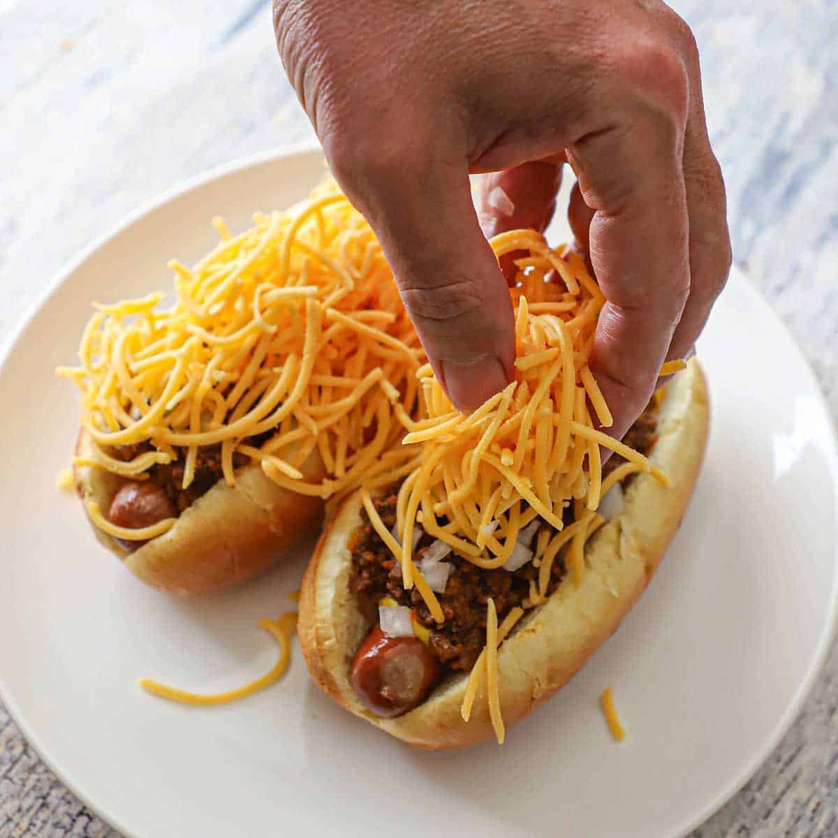 A person placing a large mound of finely shredded cheddar cheese over the tops of two Cincinnati Cheese Coney dogs with chili, onions, and mustard.