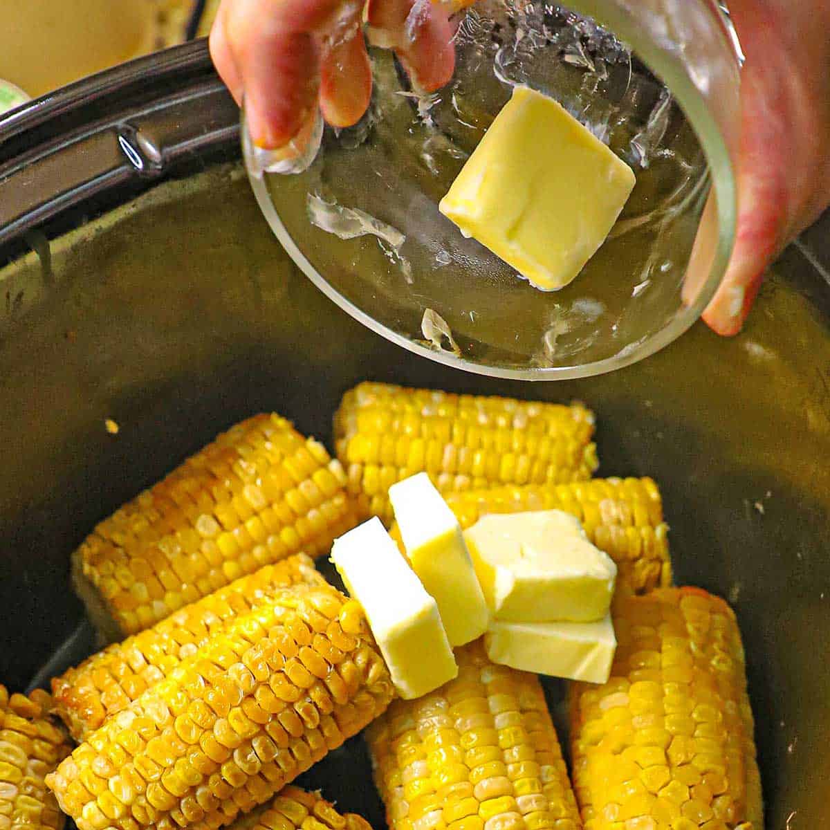 A person dumping pads of butter from a small glass bowl into the base of a black slow cooker that is filled with cooked slow cooker corn on the cob.