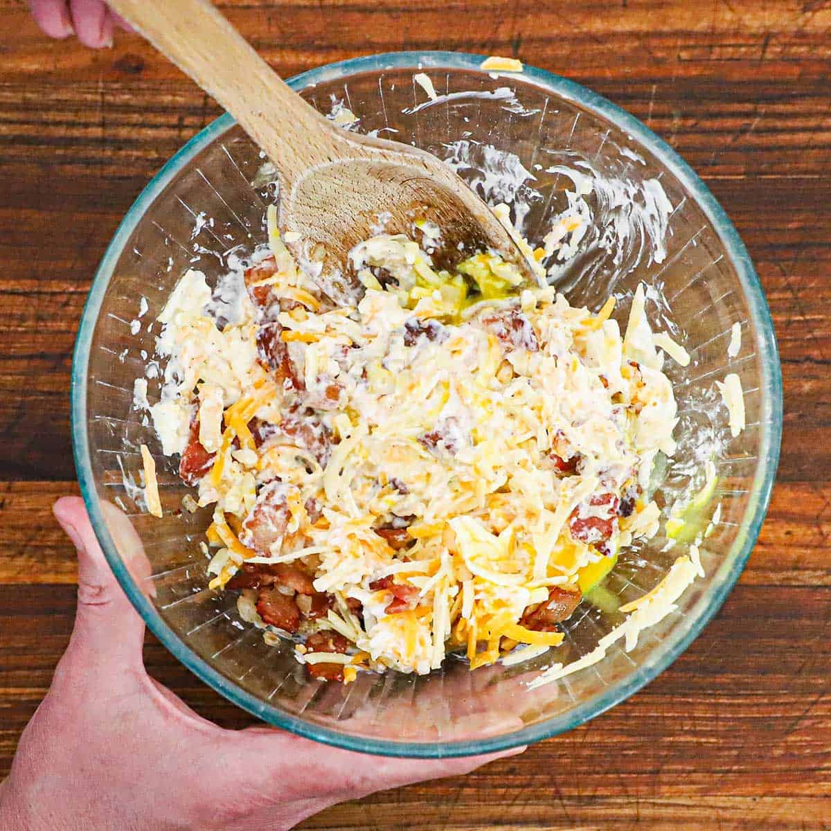A person using a large wooden spoon to stir together a mixture of shredded cheese, crispy chopped bacon, and a raw egg in a glass bowl.